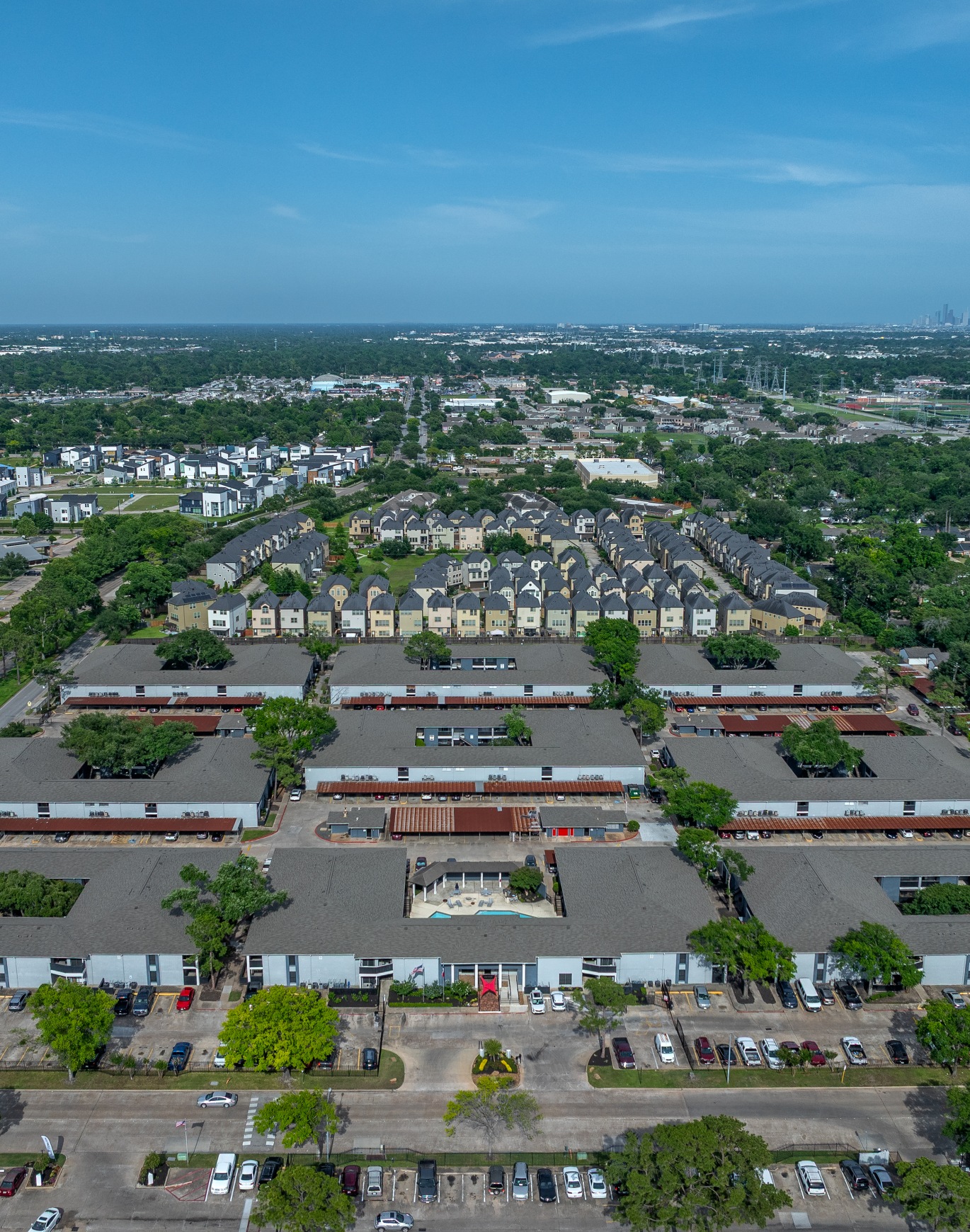 Aerial view of a residential complex with multiple buildings, parking lots, and greenery, showcasing a city skyline in the distance under a clear blue sky.